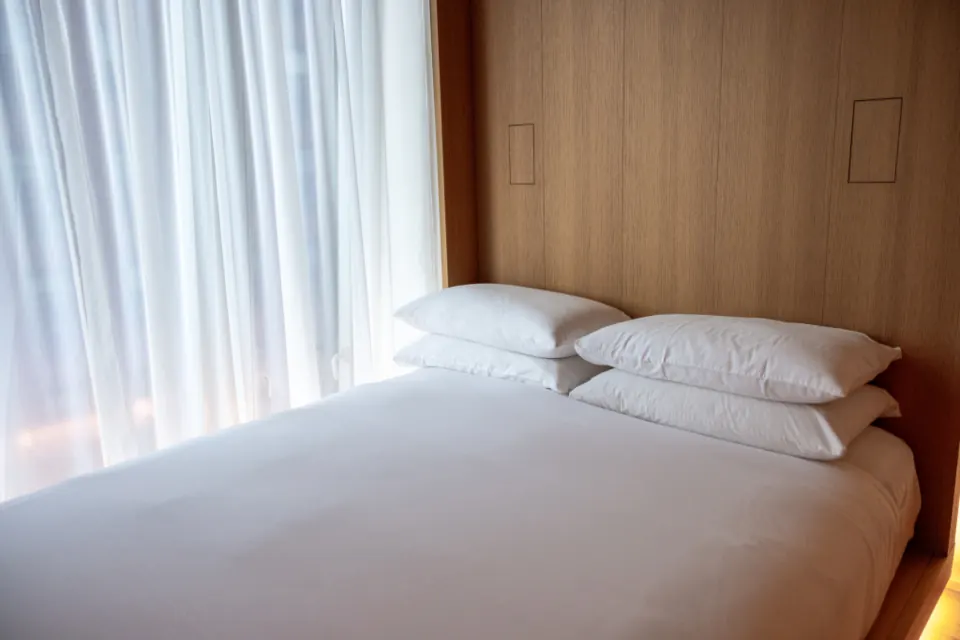 Cama con almohadas blancas y cabecero de madera junto a una ventana luminosa